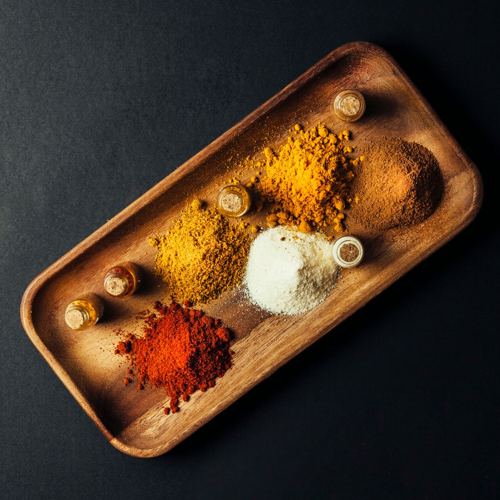 Turmeric powder, chili powder, coriander powder, and pepper powder are placed on a wooden tray against a black background.