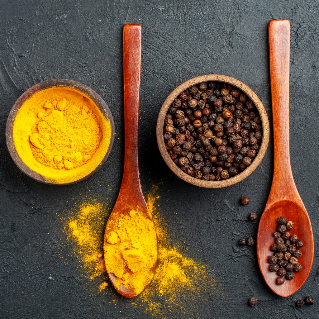 Turmeric powder and black pepper on a different wooden spoon against a black background.