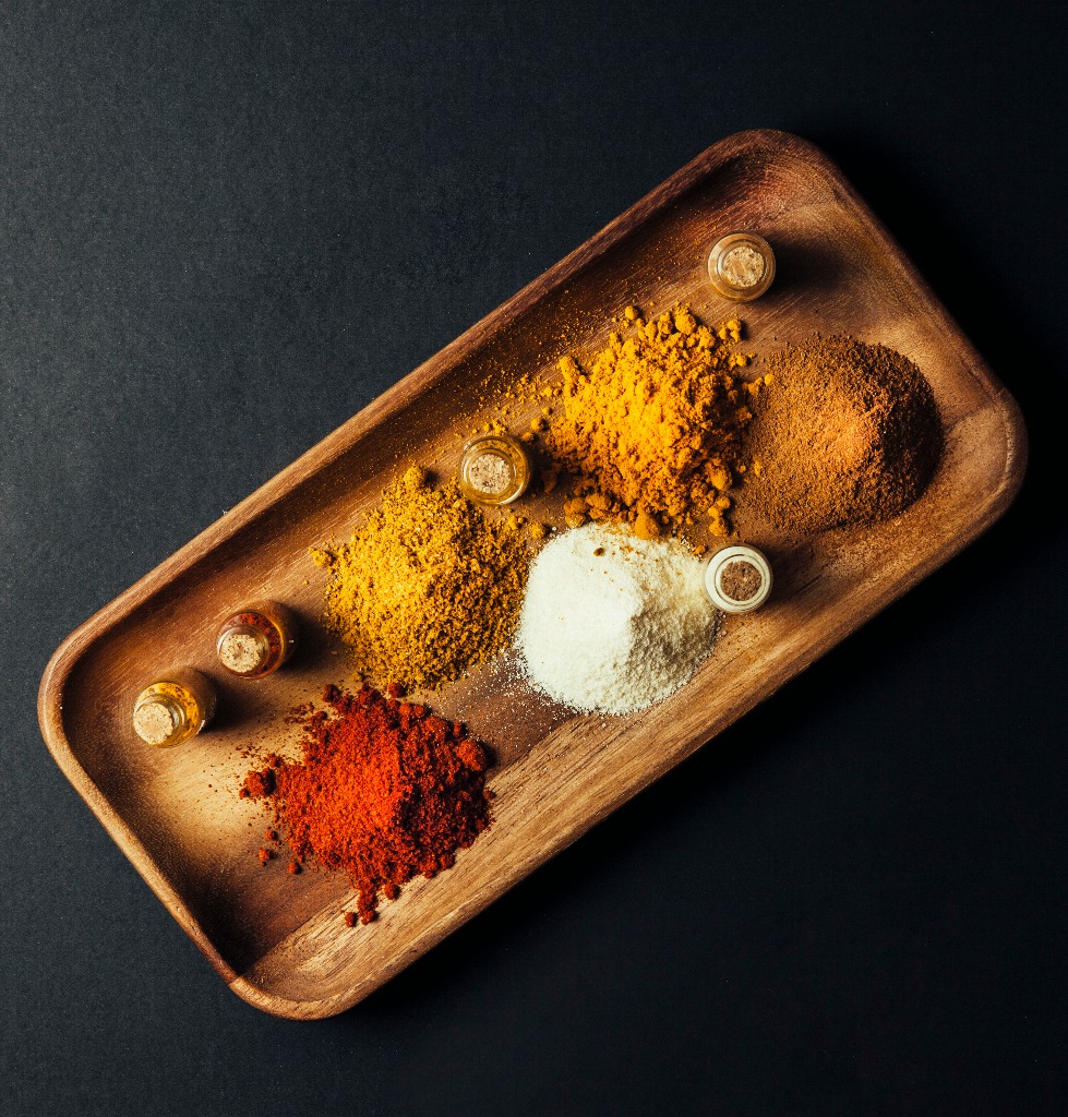 Turmeric powder, chili powder, coriander powder, and pepper powder are placed on a wooden tray against a black background.